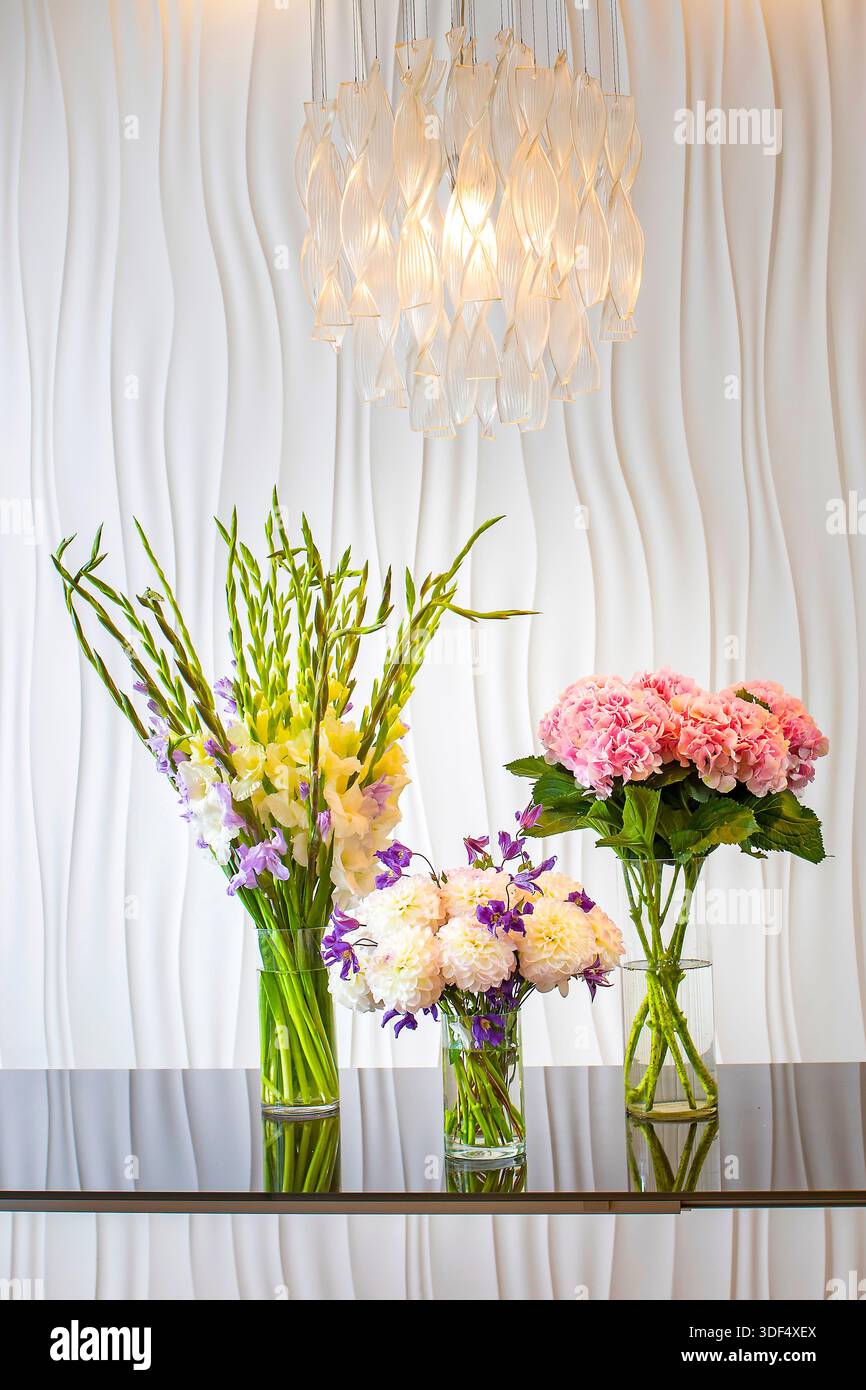 Three flower arrangements in glass vases on a black glass table against a white wavy wall, illuminated by a crystal pendant light. Stock Photo