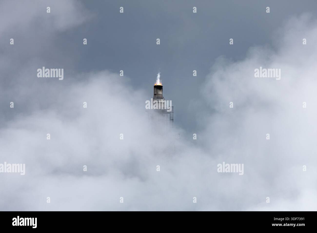 Lone flare stack shrouded in vapor clouds emits flame with sharp white plume stretching upward against background of an errie overcast gray-blue sky. Stock Photo