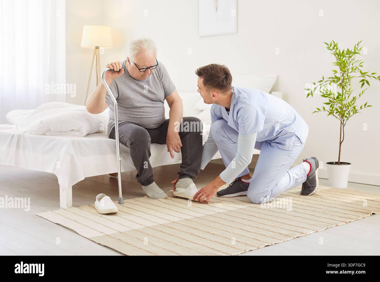 Young nurse or health care worker helping a senior patient with crutches to put on shoes at home. Stock Photo