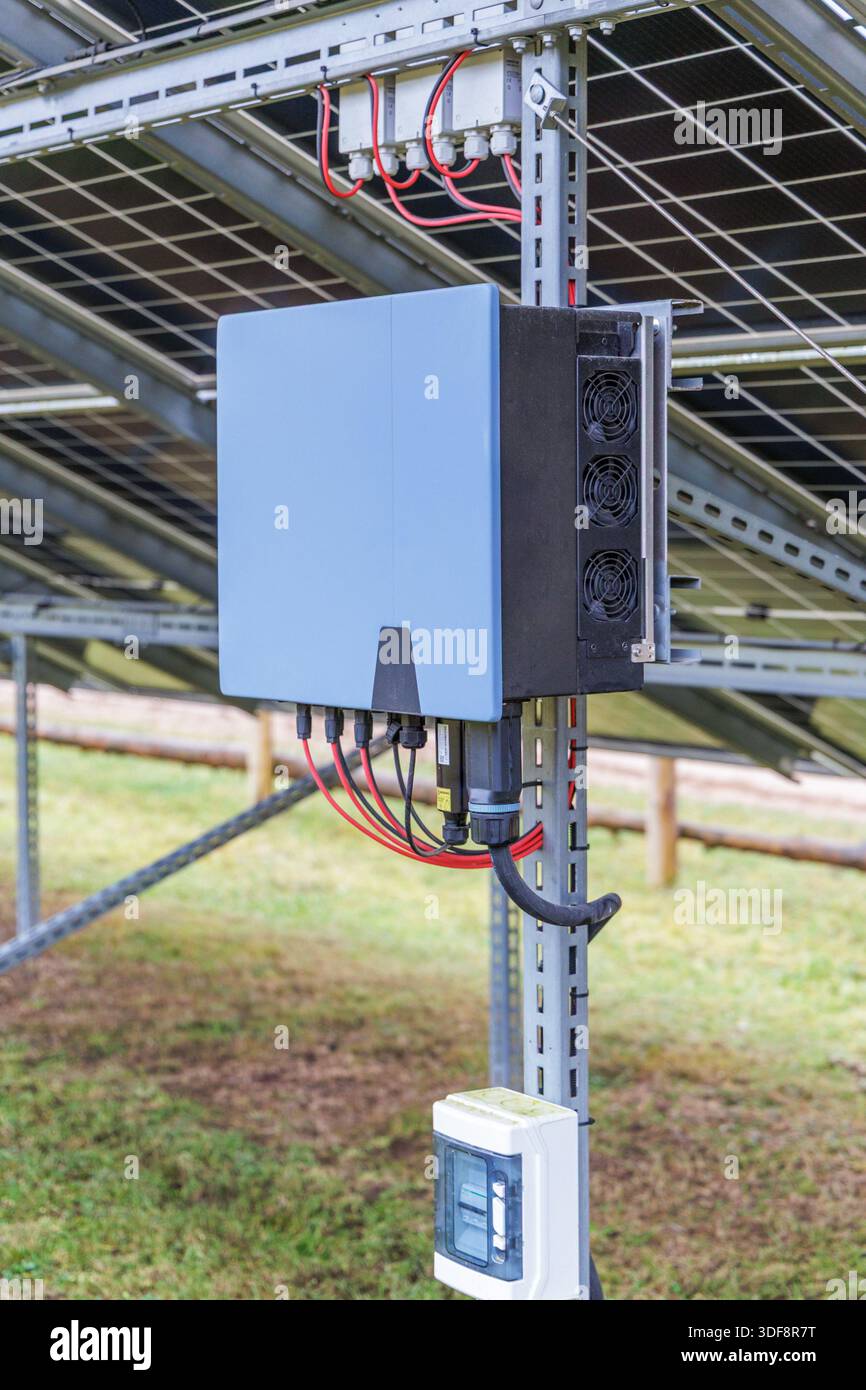 A close-up of a blue solar power inverter mounted on a metal frame under solar panels in an outdoor field. Stock Photo