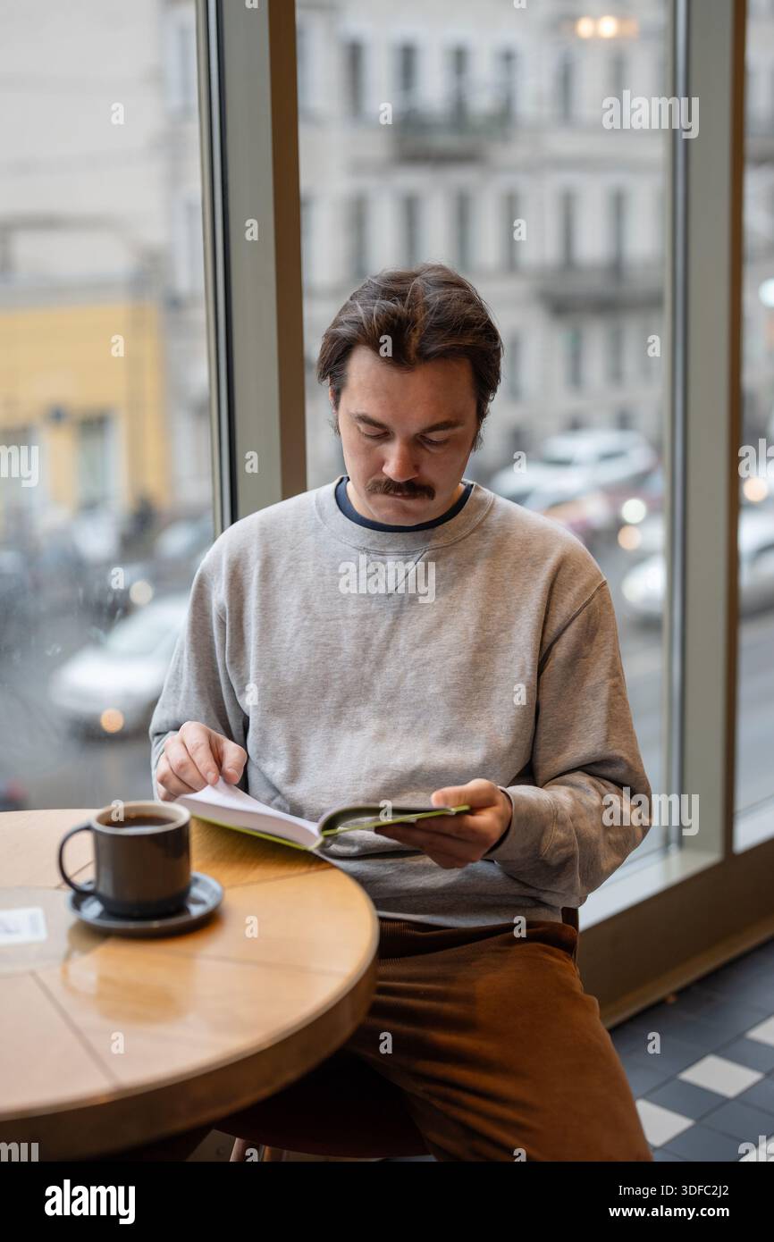 Stylish handsome man reading favorite book while sitting in cozy coffee shop during day break. Stock Photo