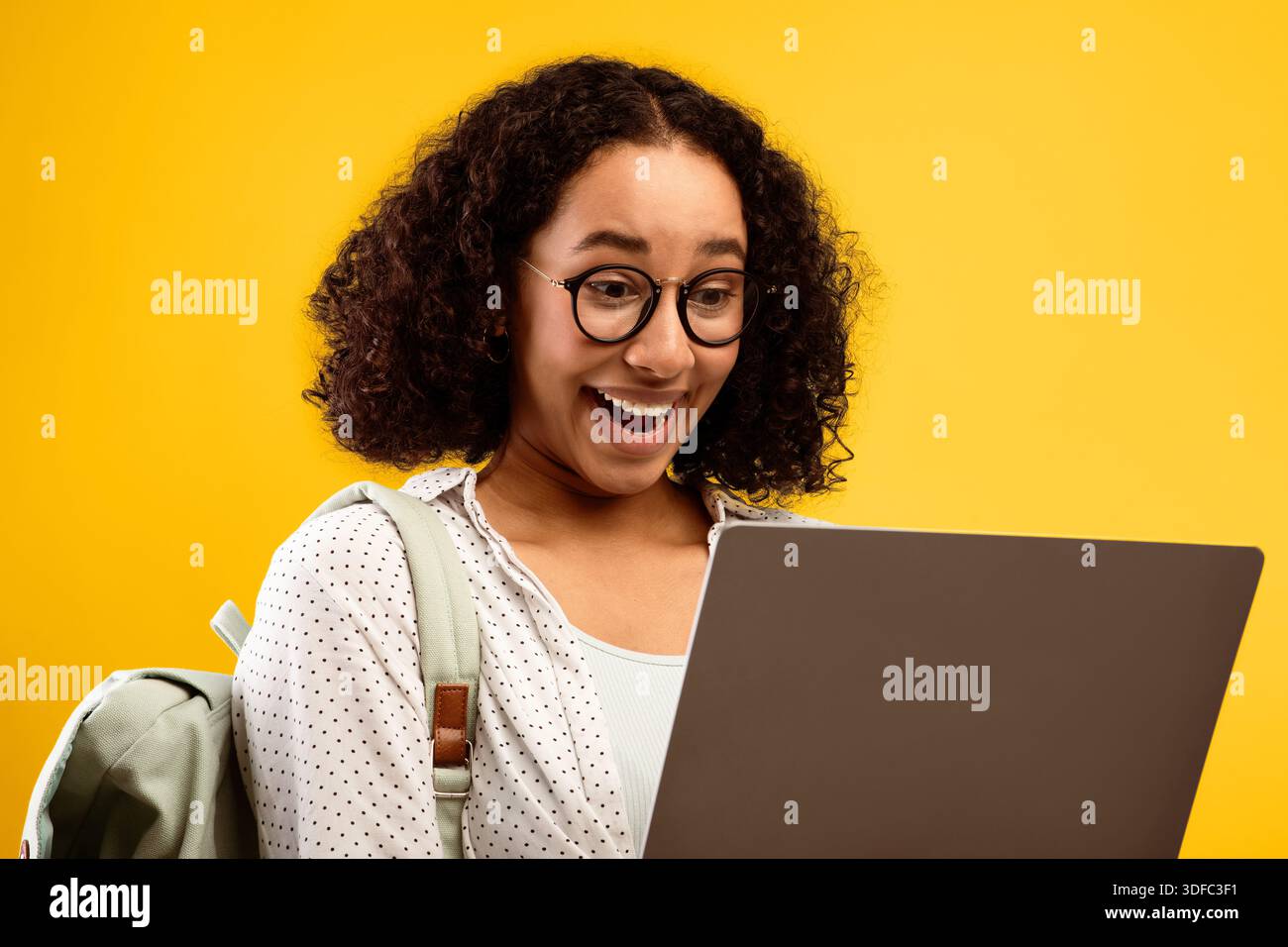 Young Woman Shows Excitement While Using Laptop With Yellow Background and Backpack Stock Photo