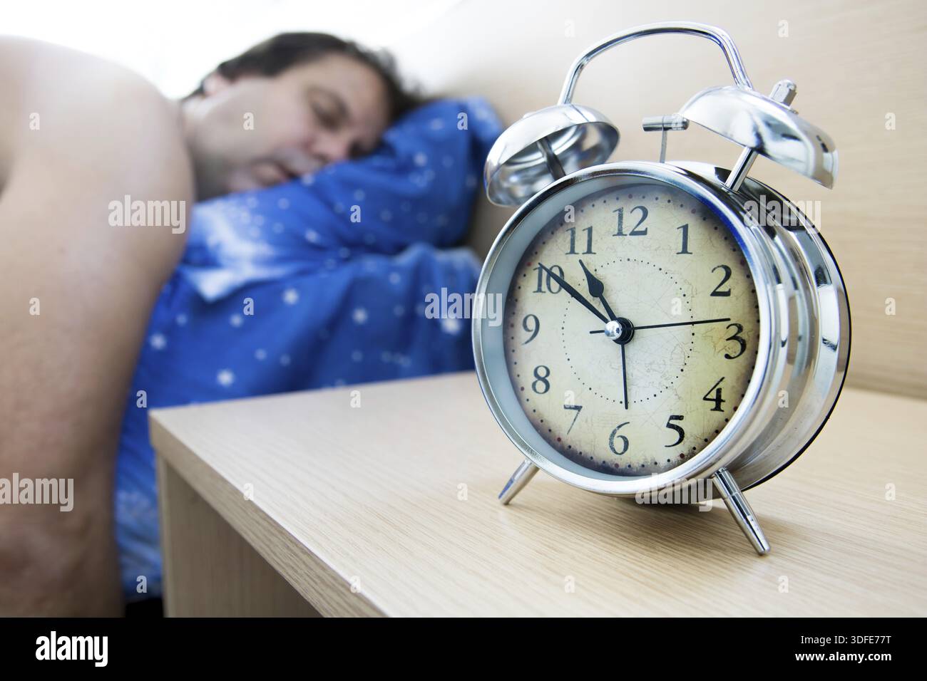 Tired man sleeping on the bed beside the alarm clock on table, in daylight. Exhausted man oversleep in the bedroom Stock Photo