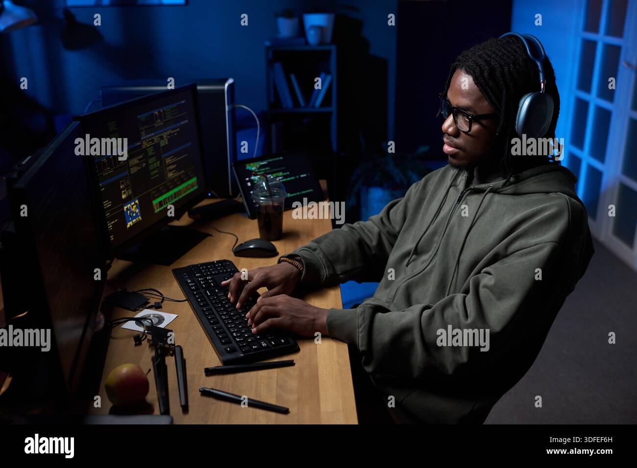 Young Black Man Wearing Headphones Coding on Computer in Dark Office Stock Photo