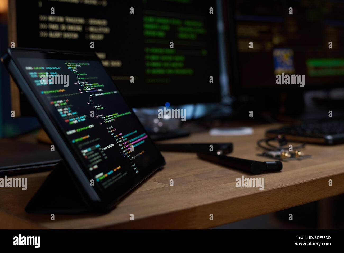 Tablet Displaying Programming Code on Desk with Computer Monitors in Background Stock Photo