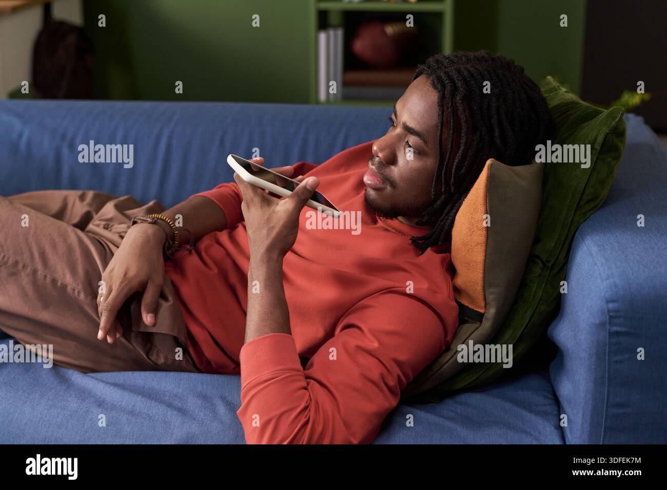 Young Black Man Relaxing on Sofa Using Smartphone Voice Assistant Stock Photo