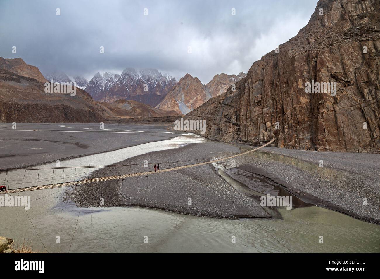View of two figures crossing a precarious rope bridge over a glacial river amidst towering, rugged mountains under a cloudy sky, Gojal, Gilgit Baltist Stock Photo