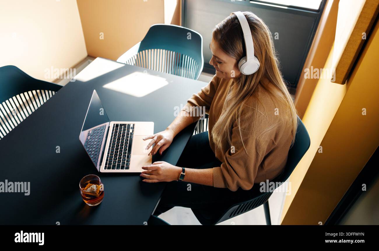 A young adult woman works on her laptop while enjoying music with headphones, creating a focused and modern setting. A relaxed and professional enviro Stock Photo