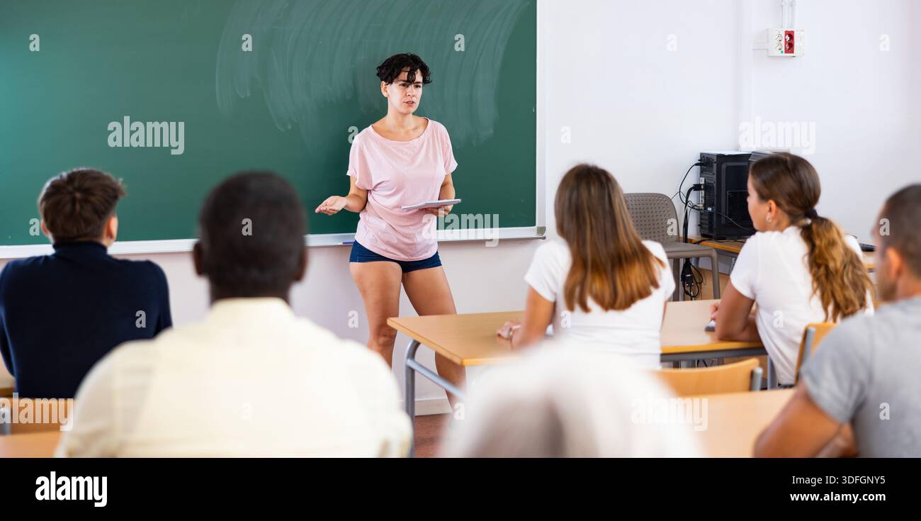 Young teacher explaining new topic while standing at blackboard Stock Photo