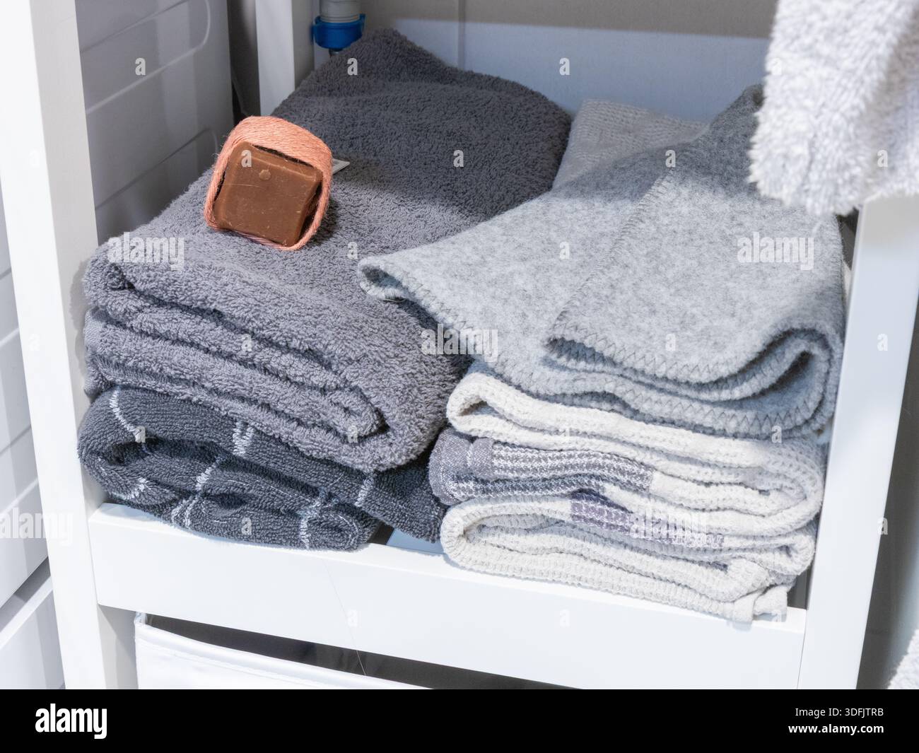 Folded grey and white towels are neatly stacked on a white shelf, with a brown, string-tied soap bar resting on the top left stack, suggesting organiz Stock Photo