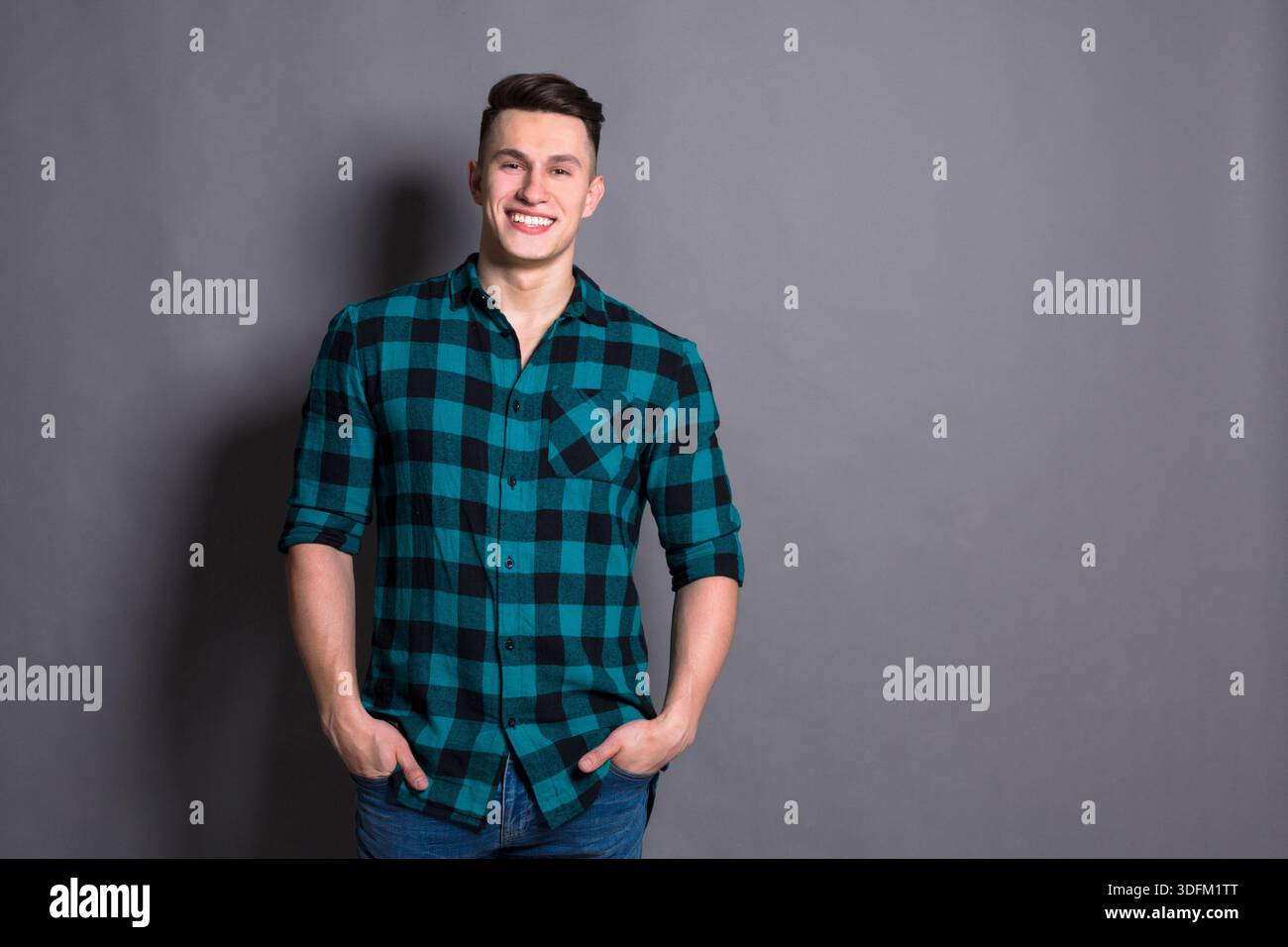 Young Man Smiling While Standing Against a Gray Backdrop With Hands in Pockets Stock Photo