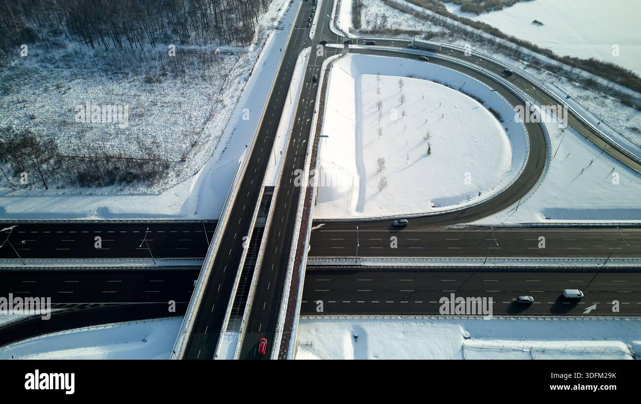 Aerial view of a multilane road interchange with curved ramps and moving vehicles, surrounded by snow-covered fields and urban infrastructure during Stock Photo