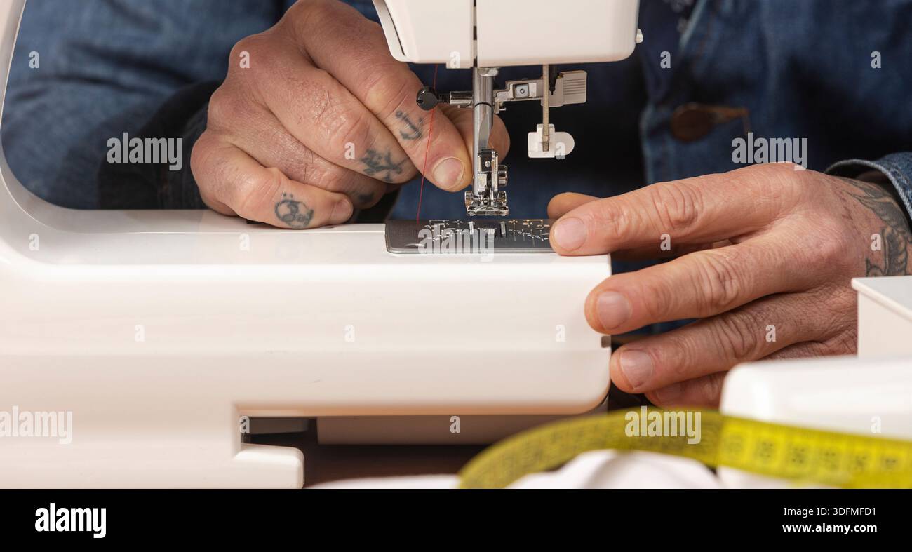 Craftsperson's tattooed hands preparing a domestic sewing machine with red thread for textile work Stock Photo