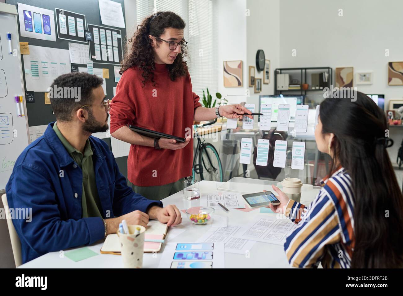 Multiethnic group of young adults collaborating on mobile app interface design, discussing wireframes and digital prototypes at creative office workspace, using tablet and printed layouts Stock Photo