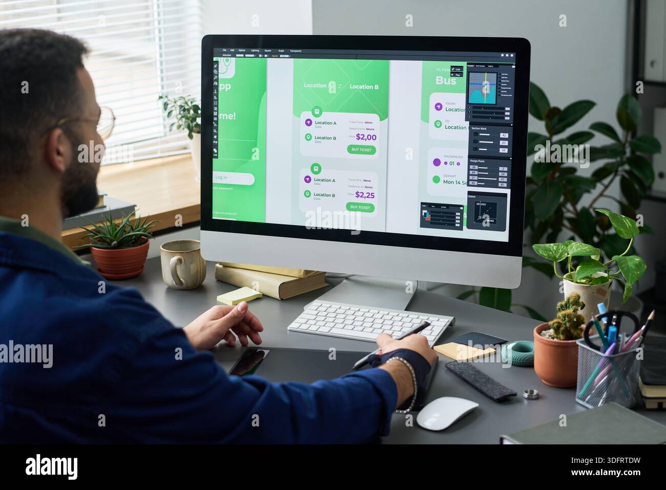 Man working on user interface design for mobile application on desktop computer, sitting at desk with digital pen and looking at large monitor displaying app screens Stock Photo
