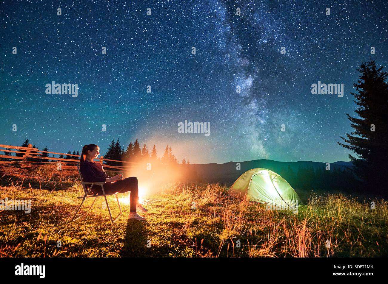 Hiker woman relaxes by campfire, illuminated by its warm glow, with tent nearby under stunning, star-filled sky. Milky Way stretches overhead, adding magical touch to serene mountain setting. Stock Photo