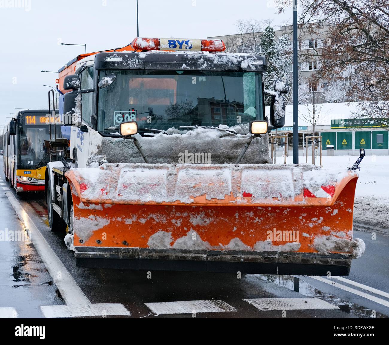 Heavy orange-bladed plow truck covered in slushy snow occupies the foreground of a wet multilane road, headlights on, articulated city bus Stock Photo