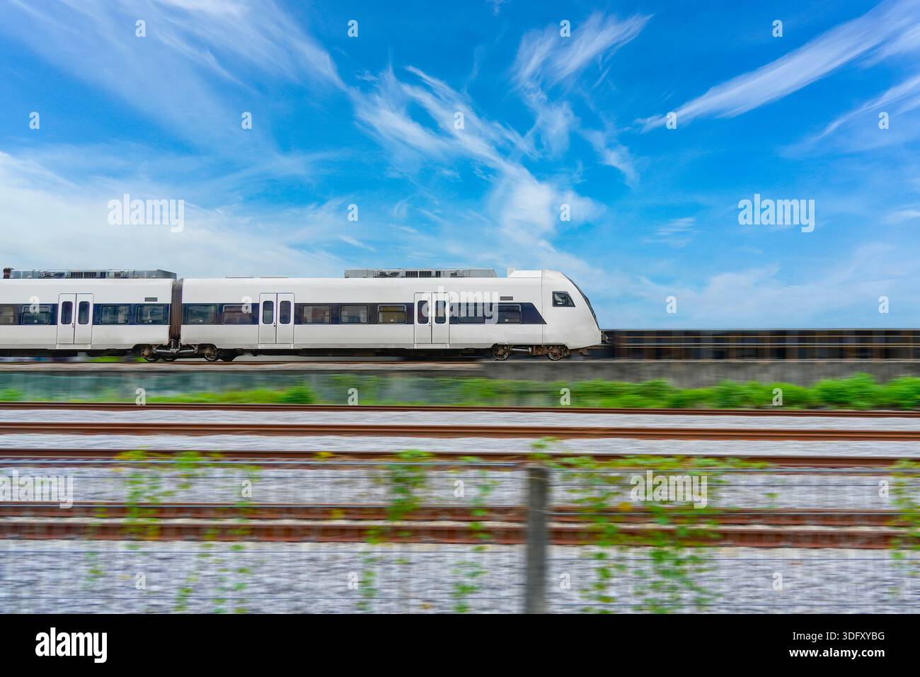 Speed modern commuter train, motion blur rides among the fields and green vegetation. with blue sky clouds, side view Stock Photo