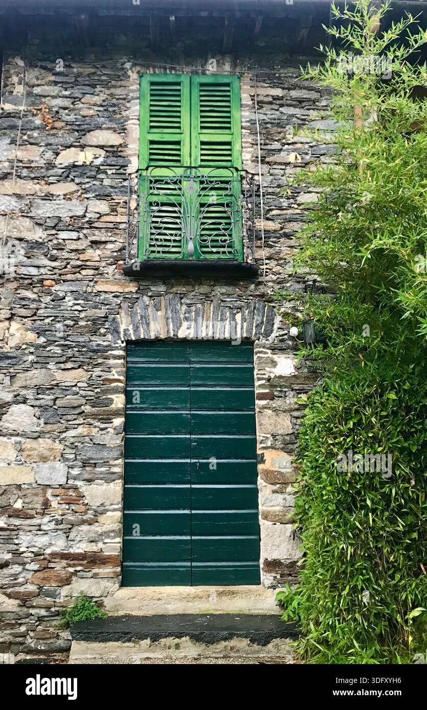 A unique house facade in Orta San Giulio, Italy, featuring a textured stone wall, green closed shutters, and a towering tree beside the building. Stock Photo