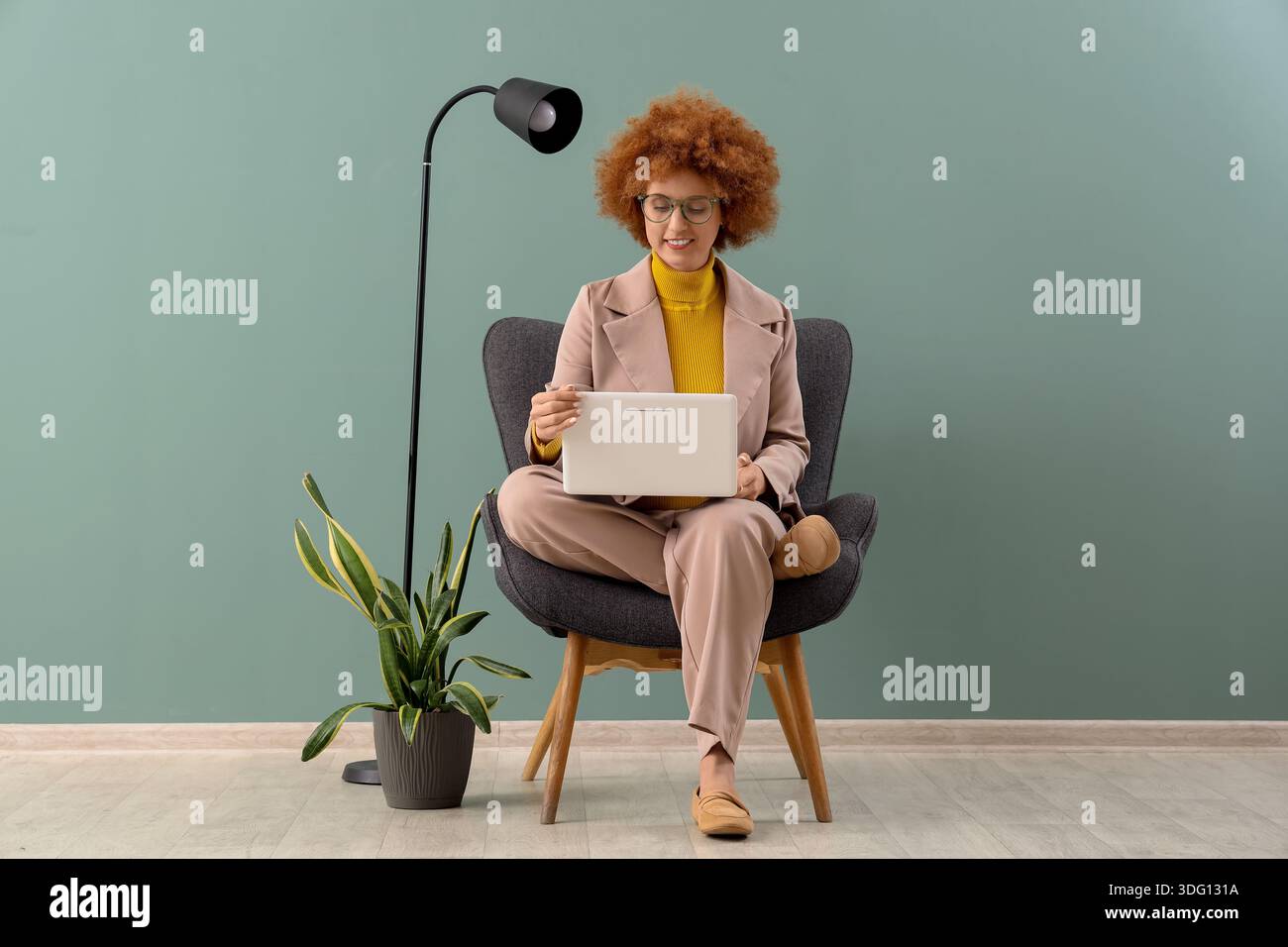Redhead female freelancer working with laptop in armchair near green wall Stock Photo