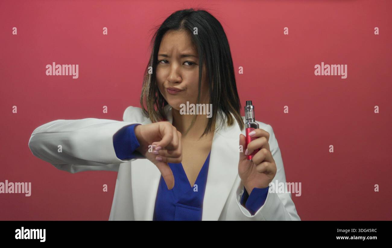Woman expressing disapproval while holding an e-cigarette against a vibrant  red background, demonstrating a negative reaction to vaping Stock Photo