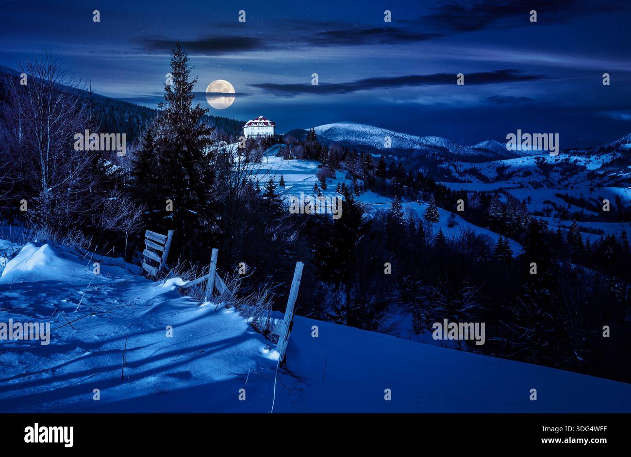 countryside in mountains at night. lovely scenery with wooden fence and coniferous forest on a snowy slopes in full moon light. village on distant hil Stock Photo
