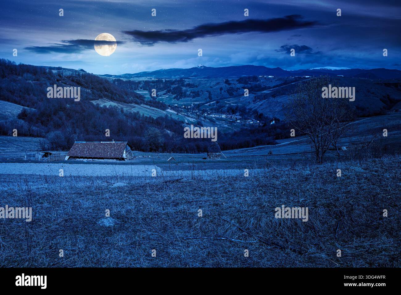 rural landscape with village in valley at night. outdoor adventures in countryside with rolling hills and snowy mountain in early spring in full moon Stock Photo