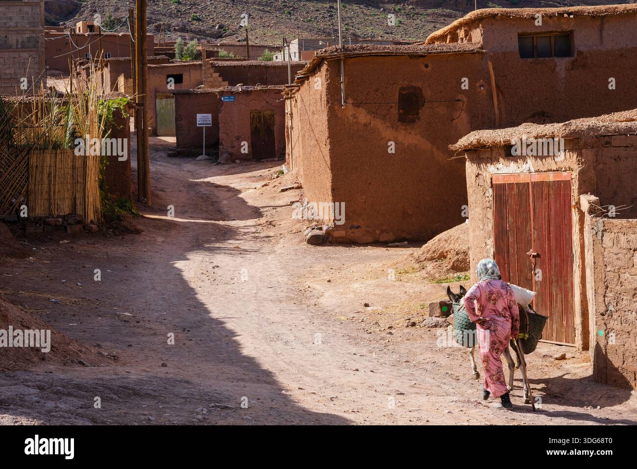 A person carrying items in a village street with rustic, earthen buildings in bright daylight. Ouarzazate, Morocco Stock Photo