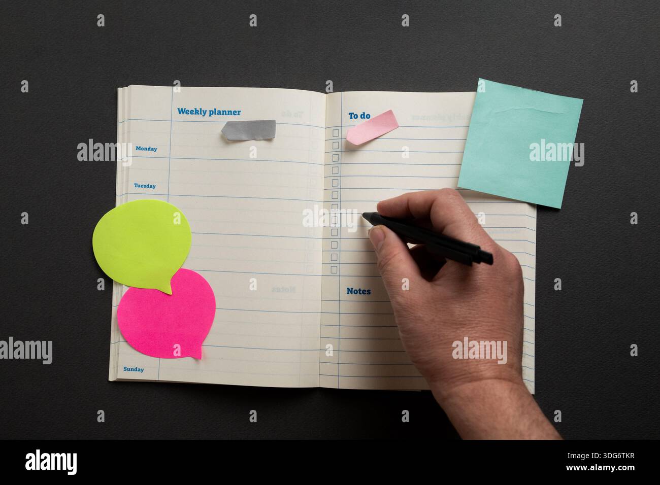 Hand writing tasks in a weekly planner with to do list, colorful sticky notes and pen on a dark desk, concept of organization, productivity and planni Stock Photo