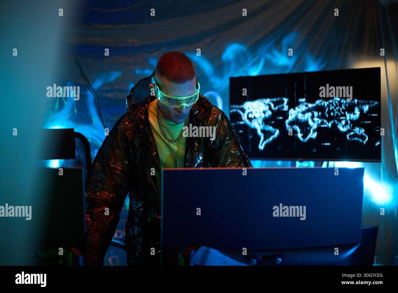 Young adult Caucasian man wearing protective glasses operating multiple computer monitors in dark room, with digital world map on screen, suggesting cybersecurity or hacking activity Stock Photo
