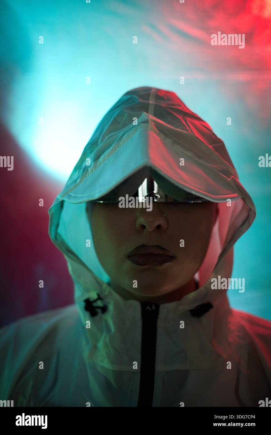 Portrait of young adult Caucasian woman wearing reflective sunglasses and hooded jacket staring directly at camera, illuminated by blue and red lights, evoking hacker theme Stock Photo