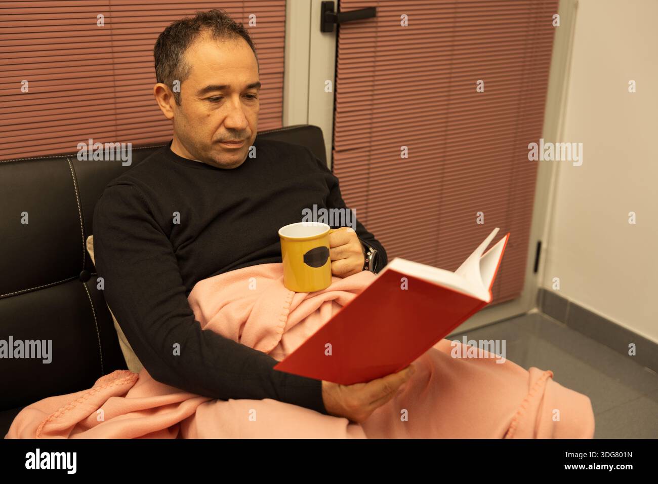 Man reading a book while resting at home with a blanket and a mug Stock Photo