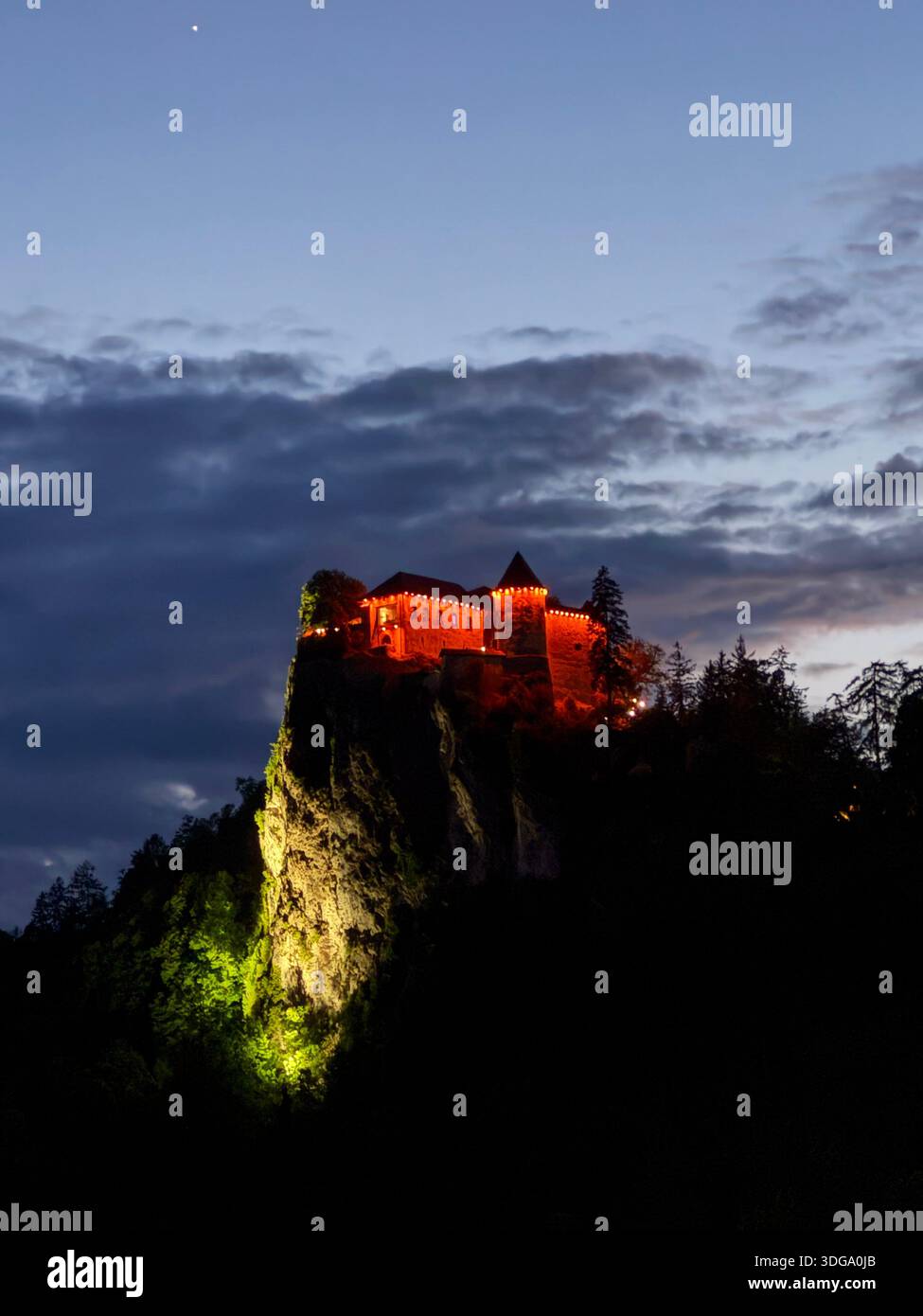 A historic cliffside landmark glows in red and gold against Slovenia’s twilight sky, framed by forest and alpine peaks - Smartphone Captured Stock Image