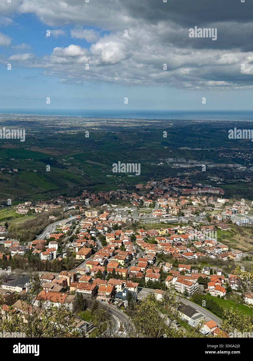 Panoramic view over an San Marino town with terracotta rooftops, rolling green hills and distant coastline under dramatic clouds - Smartphone Captured Stock Image