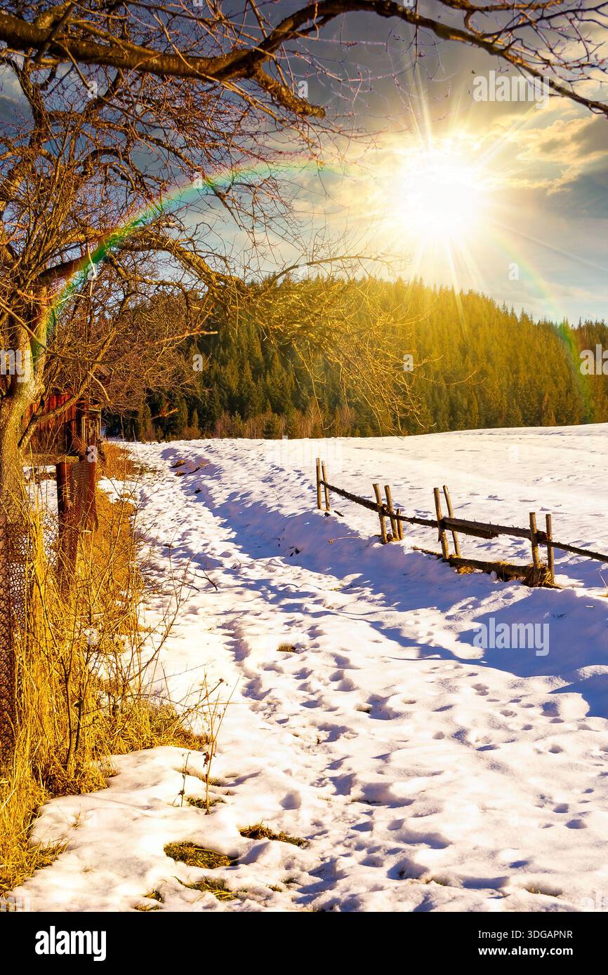 rustic wooden fence through snow covered hill at sunset. beautiful view in winter mountains in evening light. alpine rural landscape near forest. lead Stock Photo