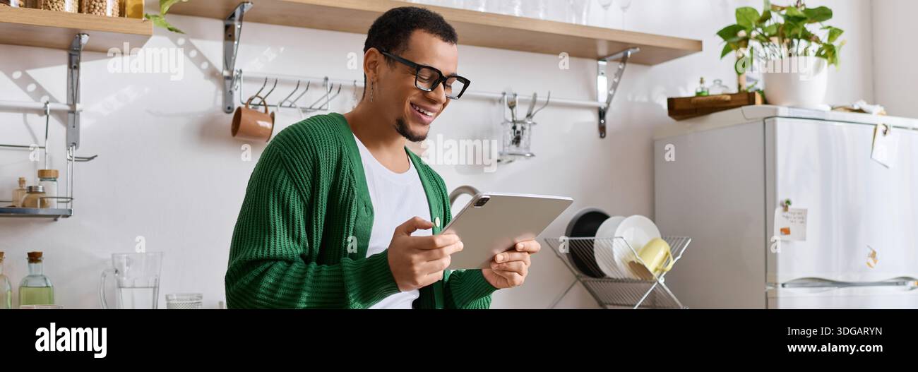 A stylish young man smiles while using a tablet in a warm and inviting kitchen atmosphere. Stock Photo