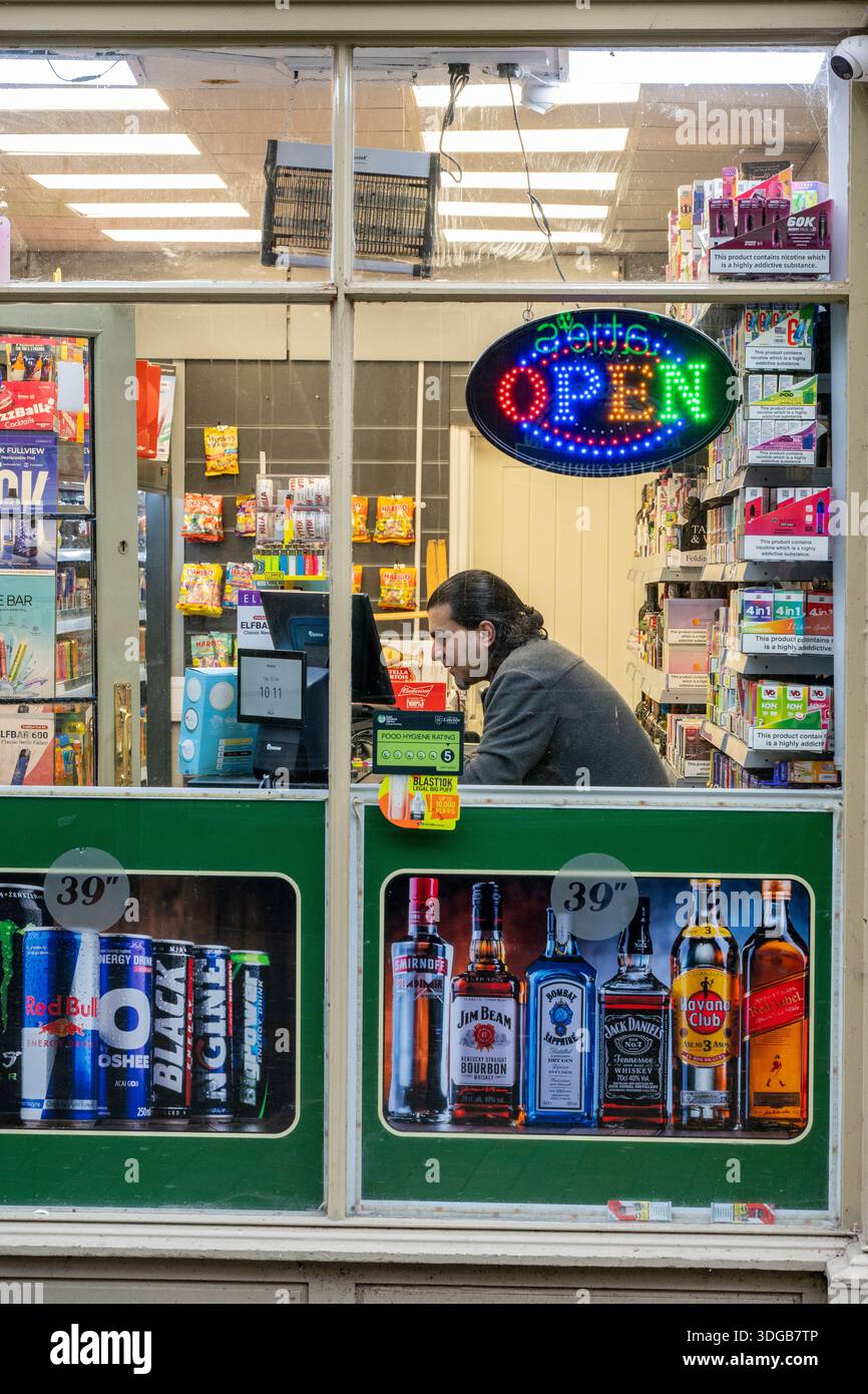 Colourful convenience store selling vapes and alcohol with young male  shopkeeper sitting behind the counter using his mobile phone or smartphone  Stock Photo