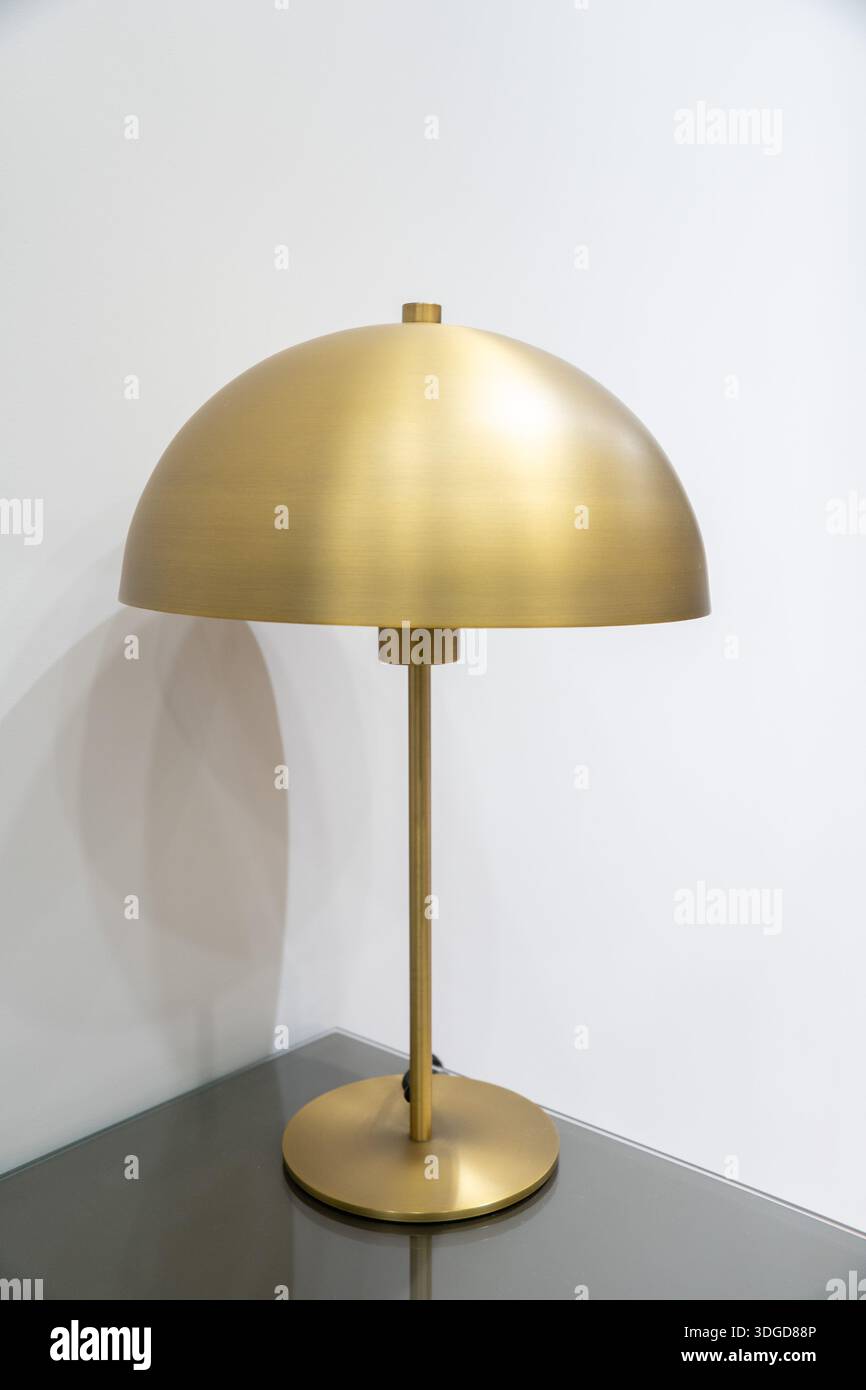 Modern gold metal table lamp with dome-shaped shade standing on a bedside table against a white wall. Suitable for concepts of interior design Stock Photo