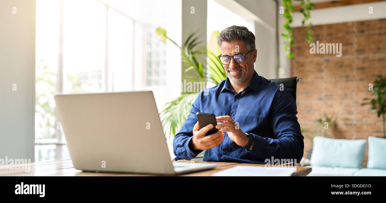 Happy middle aged businessman using mobile phone working in office. Stock Photo