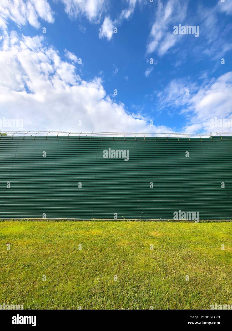 A long, dark green corrugated metal wall stretches horizontally above a strip of green grass. - Smartphone Captured Stock Image
