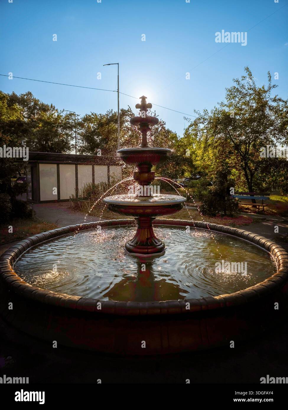 Kyiv, Ukraine. A multi-tiered fountain sprays water in a small park at Revutsky Street 7, located behind a bus stop, surrounded by trees. - Smartphone Captured Stock Image