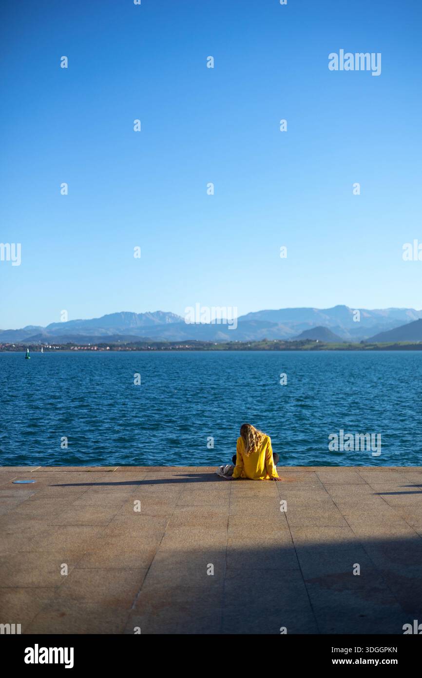 Santander, Spain – Nov 19, 2021: A woman in a yellow jacket sits alone on the waterfront promenade in Santander, looking out at the bay and distant mo Stock Photo