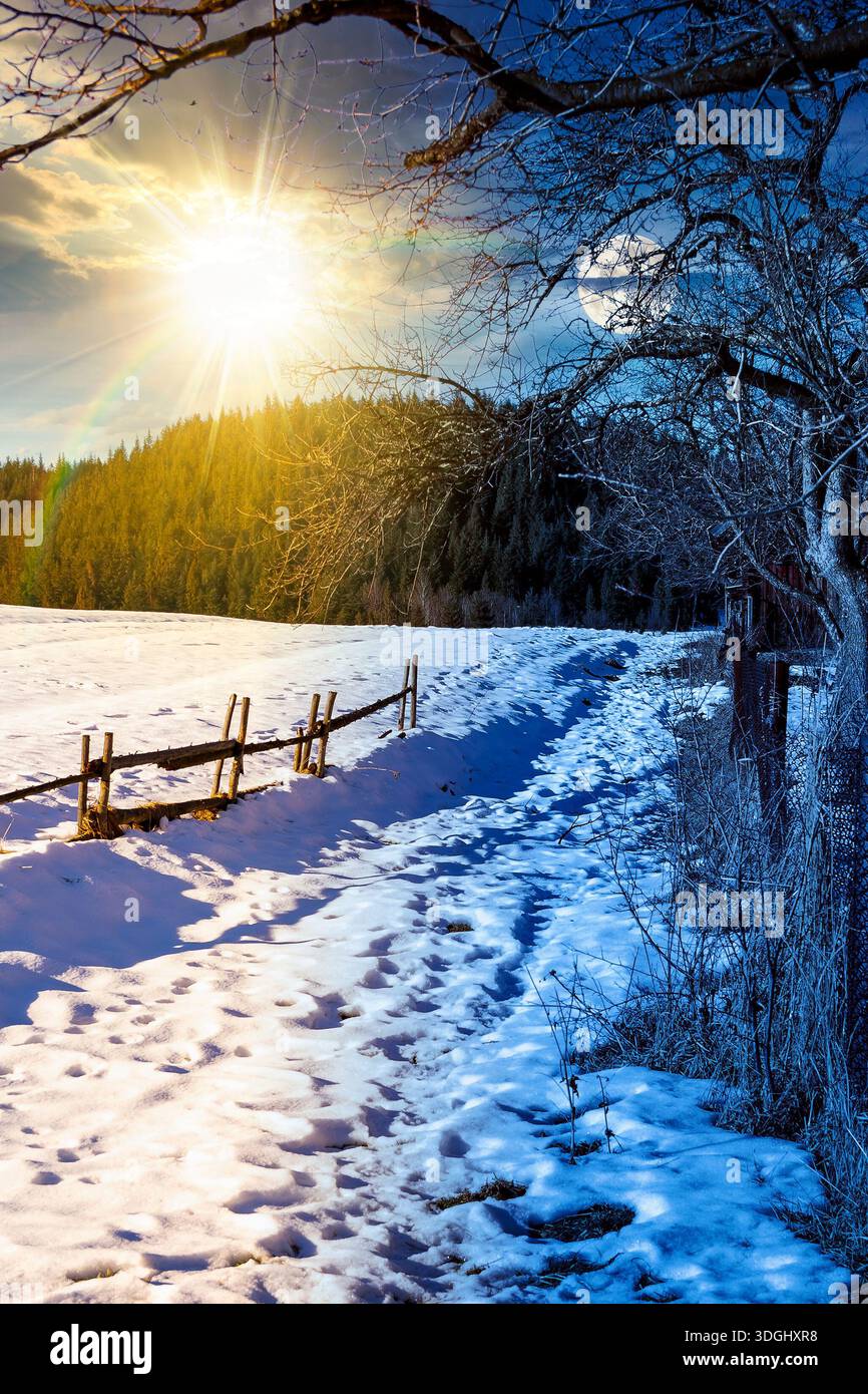 rustic wooden fence through snow covered hill. day and night time change concept. beautiful view in winter mountains with sun and moon. alpine rural l Stock Photo