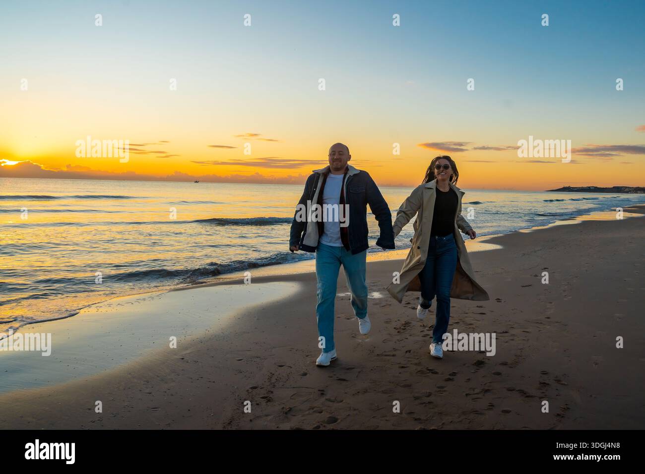 Beach sunset walk, Energized pair strolling on sandy coast at dusk, Couple taking purposeful steps on shoreline during vibrant sunset Stock Photo