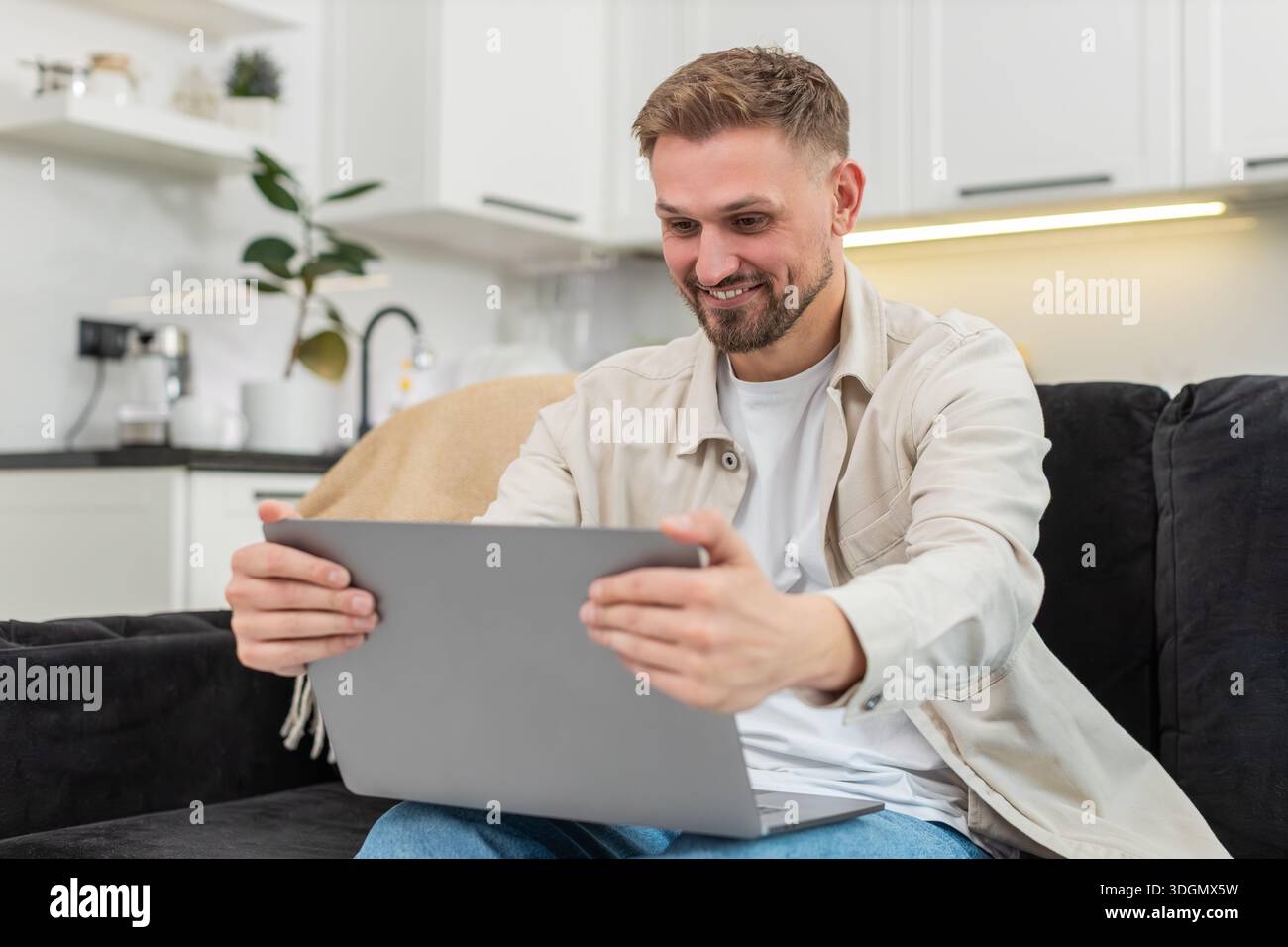 Young man at home works as freelancer checks laptop sees project approval and celebrates with joy. Caucasian guy on sofa raises hand shows victory ges Stock Photo
