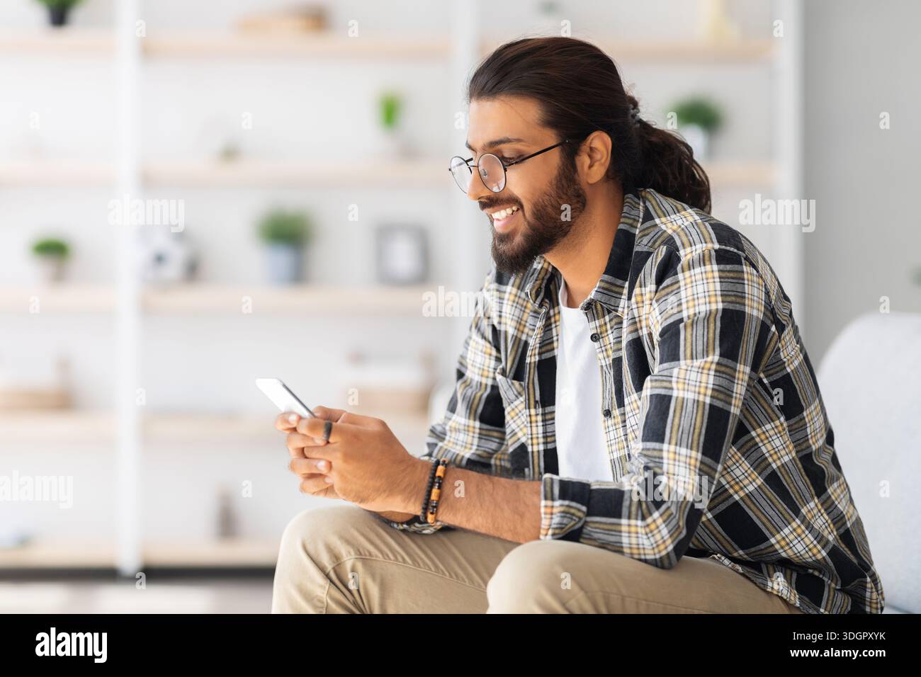 Positive long-haired middle-eastern guy sitting on couch, using cellphone Stock Photo