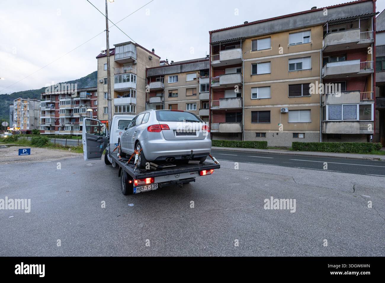 BUGOJNO, BIH, 05.23.2025:A silver sedan is loaded onto a flatbed tow truck on a city street beside a row of apartment buildings. Urban, calm atmospher Stock Photo