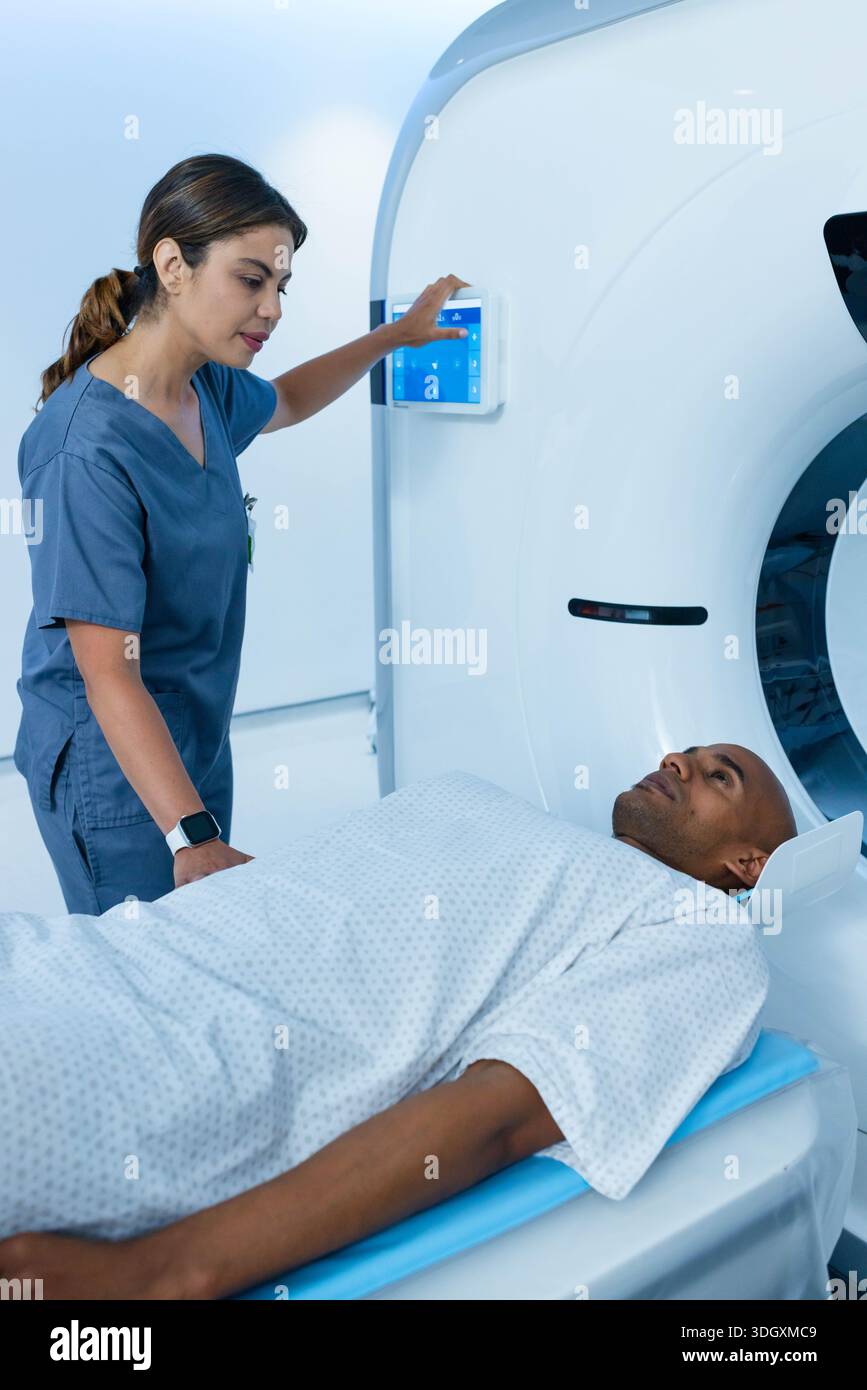 Female nurse wearing scrubs operating panel while male patient lying on CT table in gown. Scanner, radiology, hospital, touchscreen, clinical, sterile Stock Photo