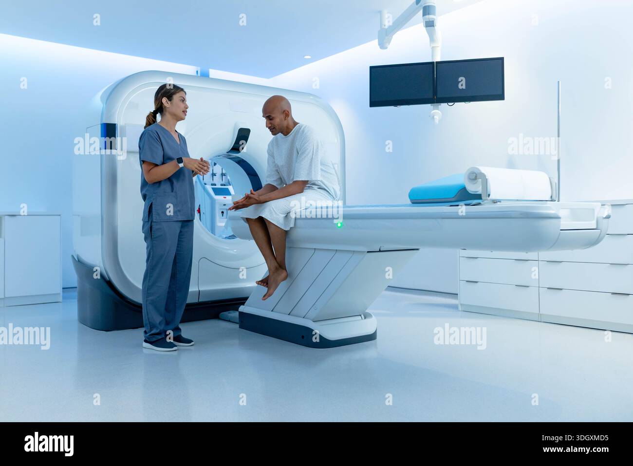 African American patient sitting on MRI table in gown, tech in scrubs speaking near gantry monitors. Radiology, imaging, clinical, hospital, diagnosti Stock Photo