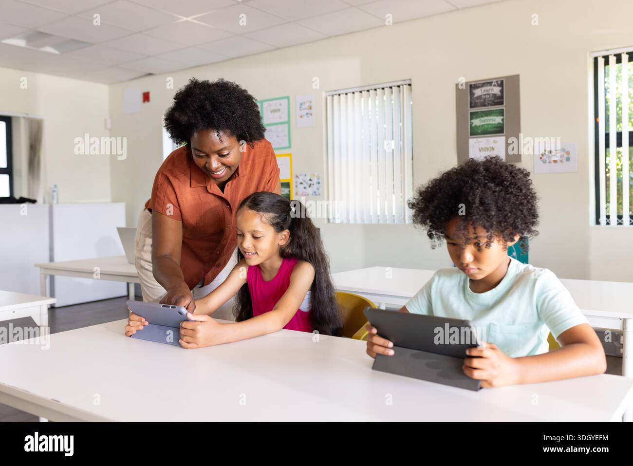 Diverse educator in rust-orange blouse guiding two youth using tablets at white table in classroom Stock Photo